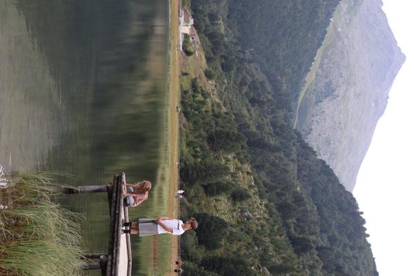 Two individuals stand on a rustic wooden pier, gazing out over a tranquil mountain lake
